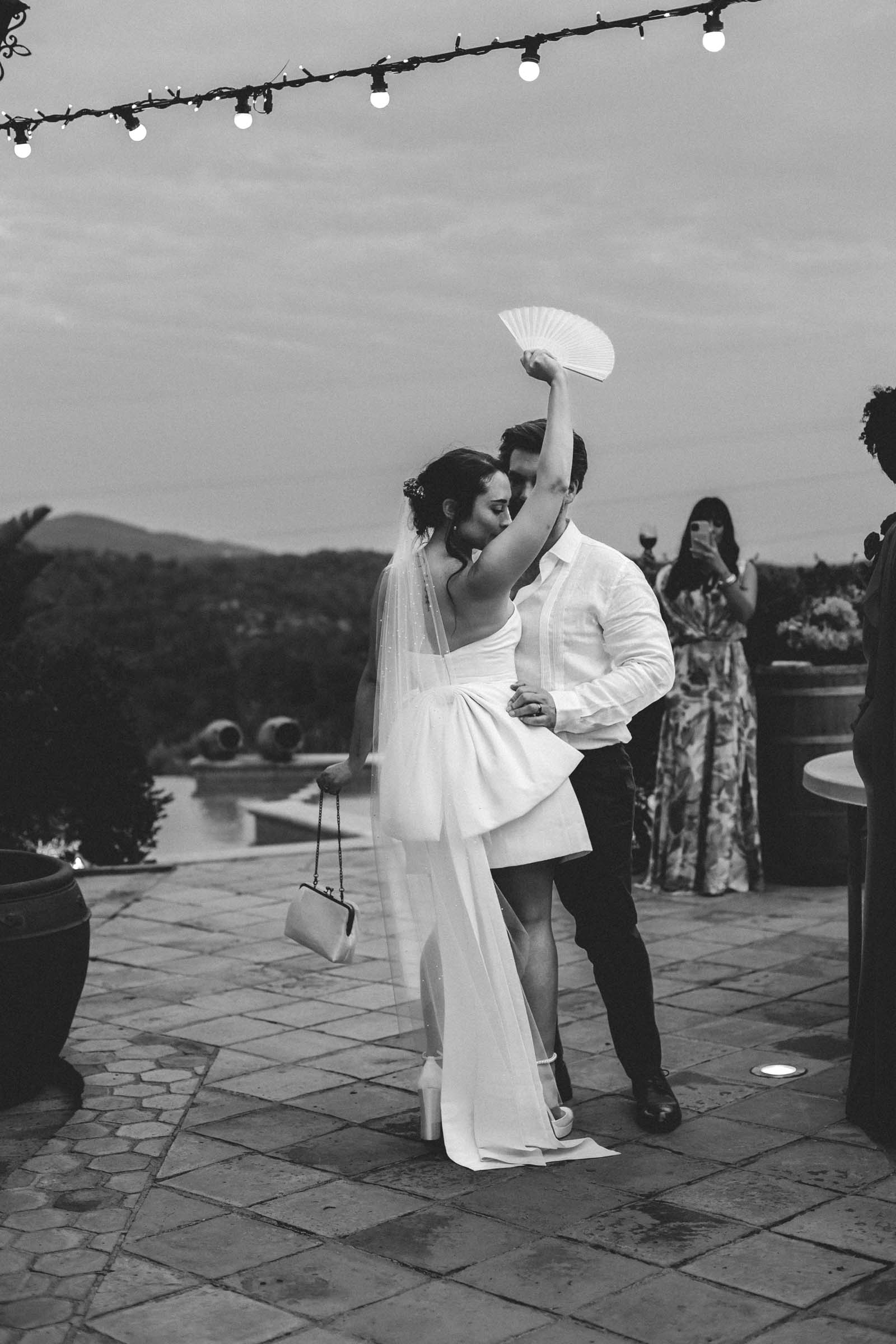 Bride wearing a short mini white dress with giant floor length bow on the back holding a white paper fan and small handbag