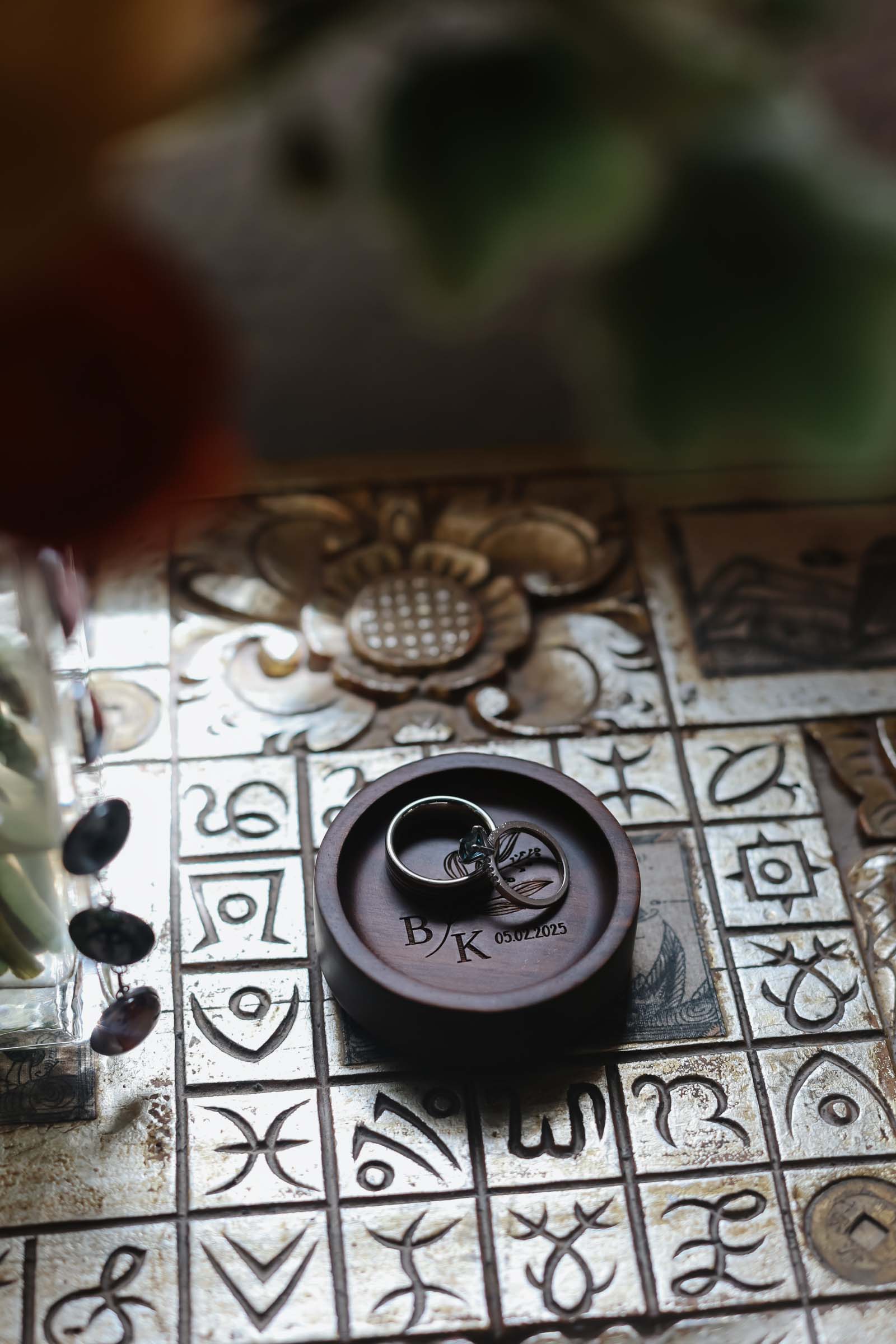 A wooden wedding ring holder with engraved initials and two rings on an ornate Spanish tile table.