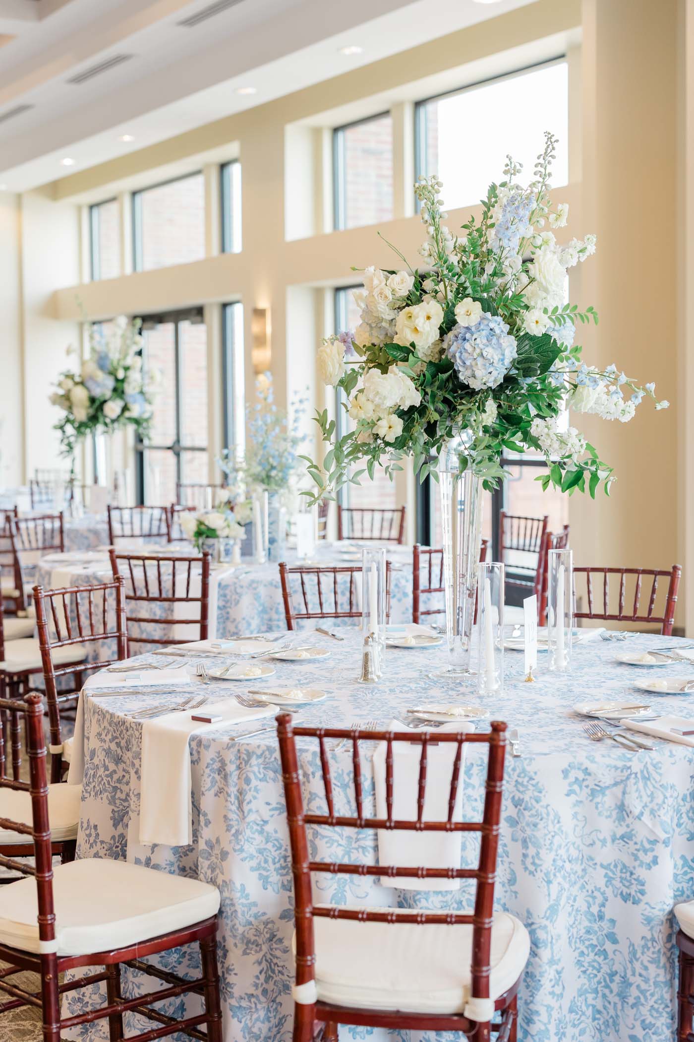 Tall white and blue wedding floral centerpieces in glass vases on a patterned blue table linen