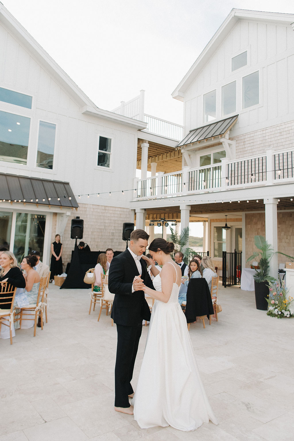Barefoot bride and groom sharing their first dance under bistro lights at their beach house wedding