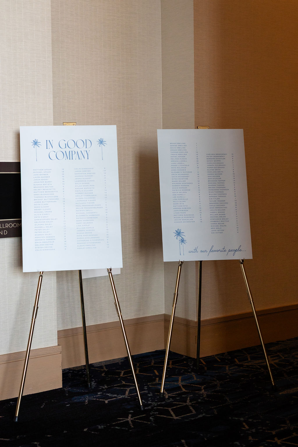 Contemporary wedding seating chart with palm trees and In Good Company heading on gold easel stands